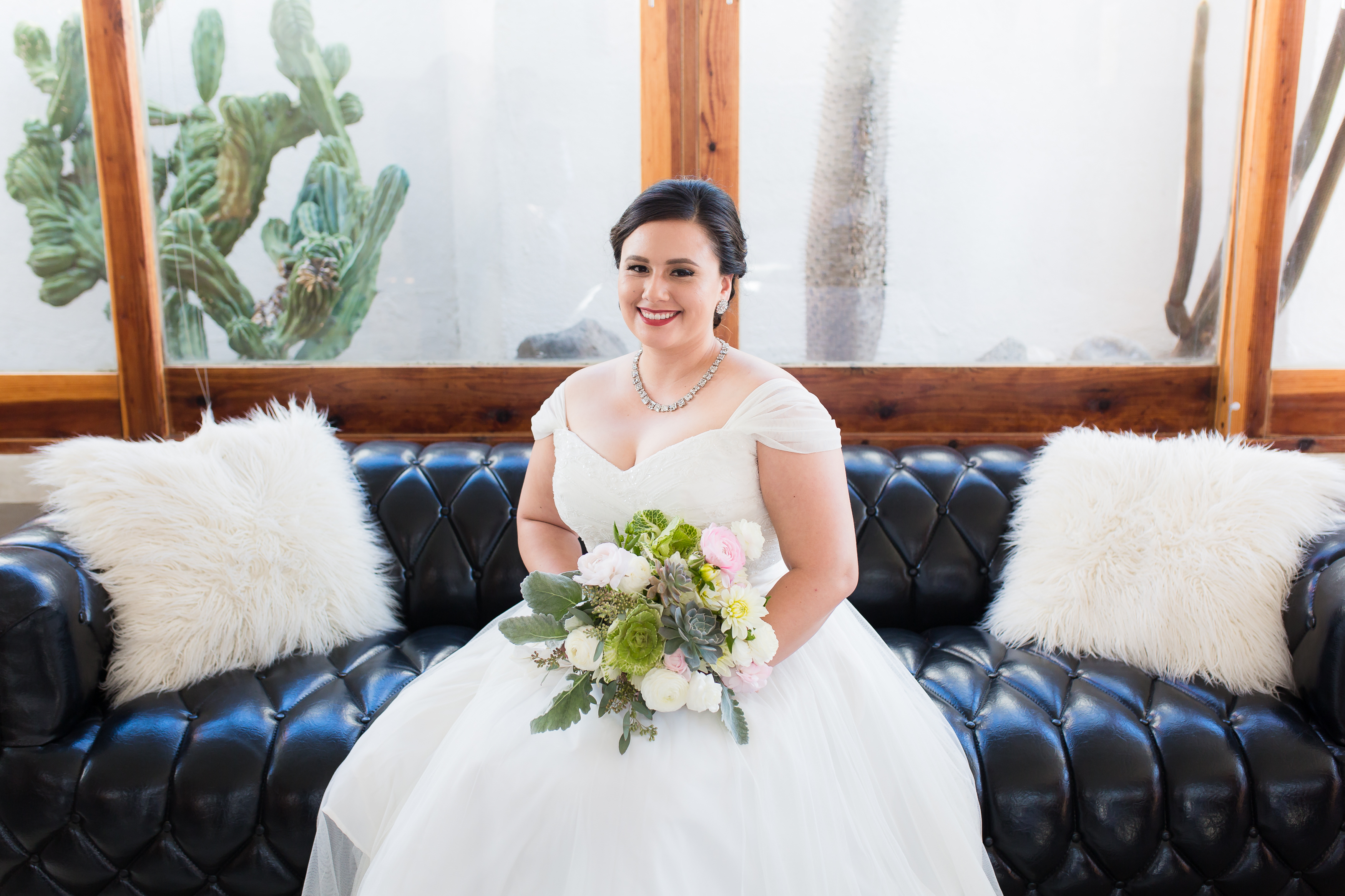Brunette bride in off the shoulder wedding dress sitting on black leather couch