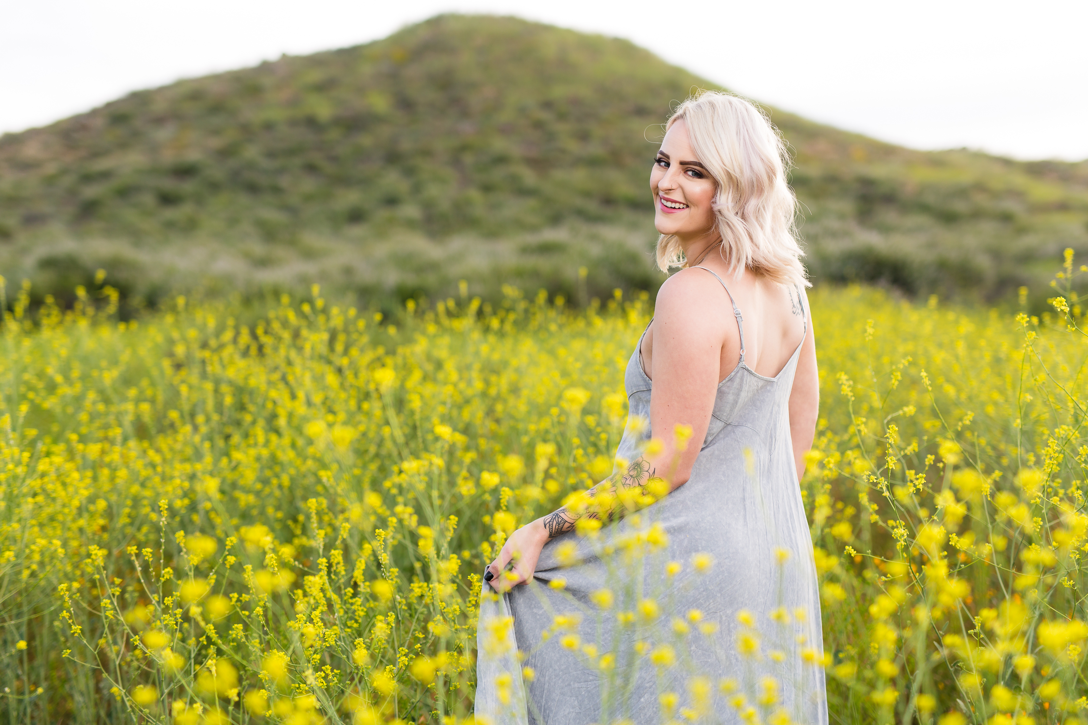 Woman walking through field of flowers towards green hill looking back and smiling
