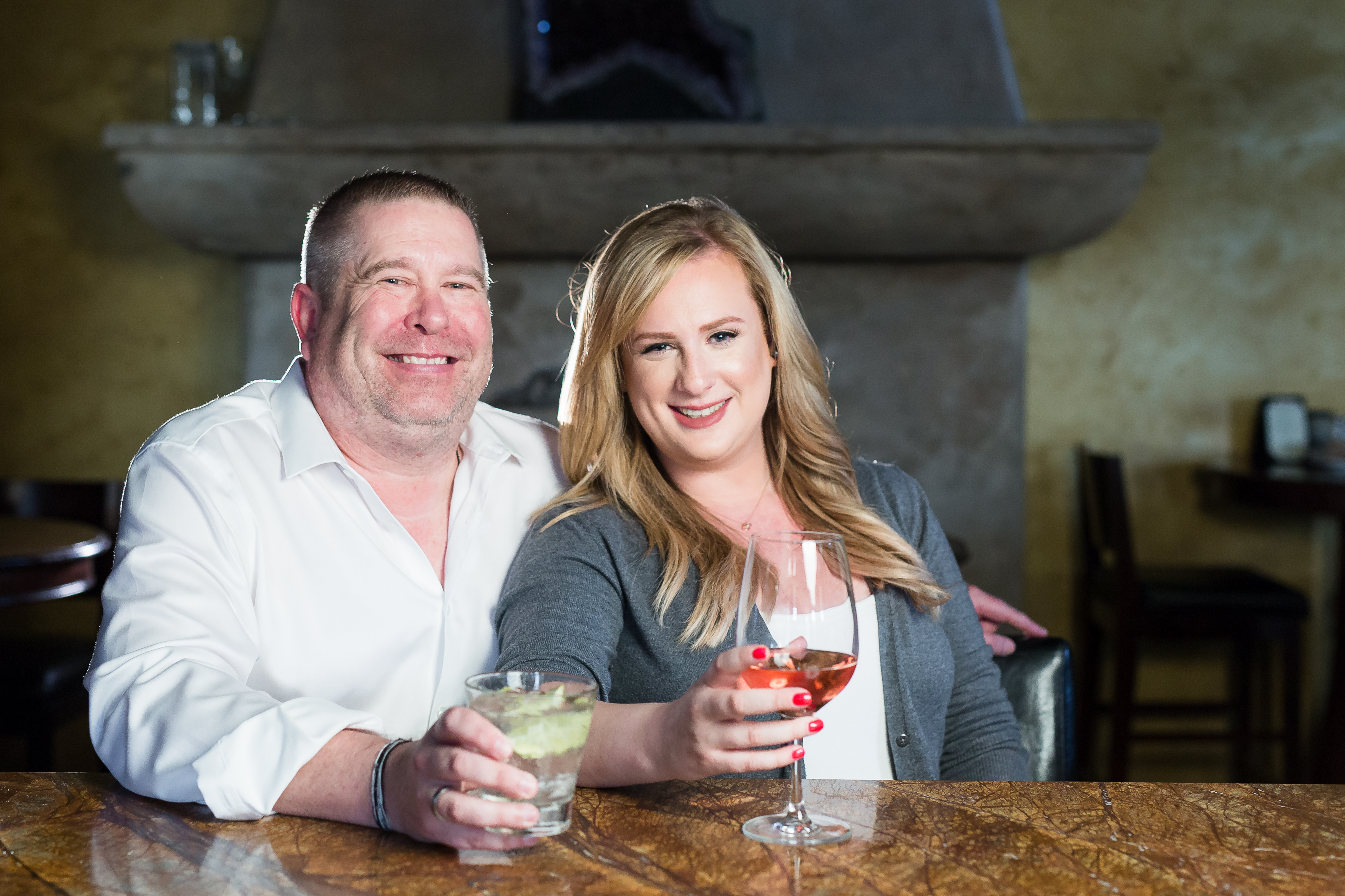 Couple smiling at the camera with alcohol in hand while sitting at the bar