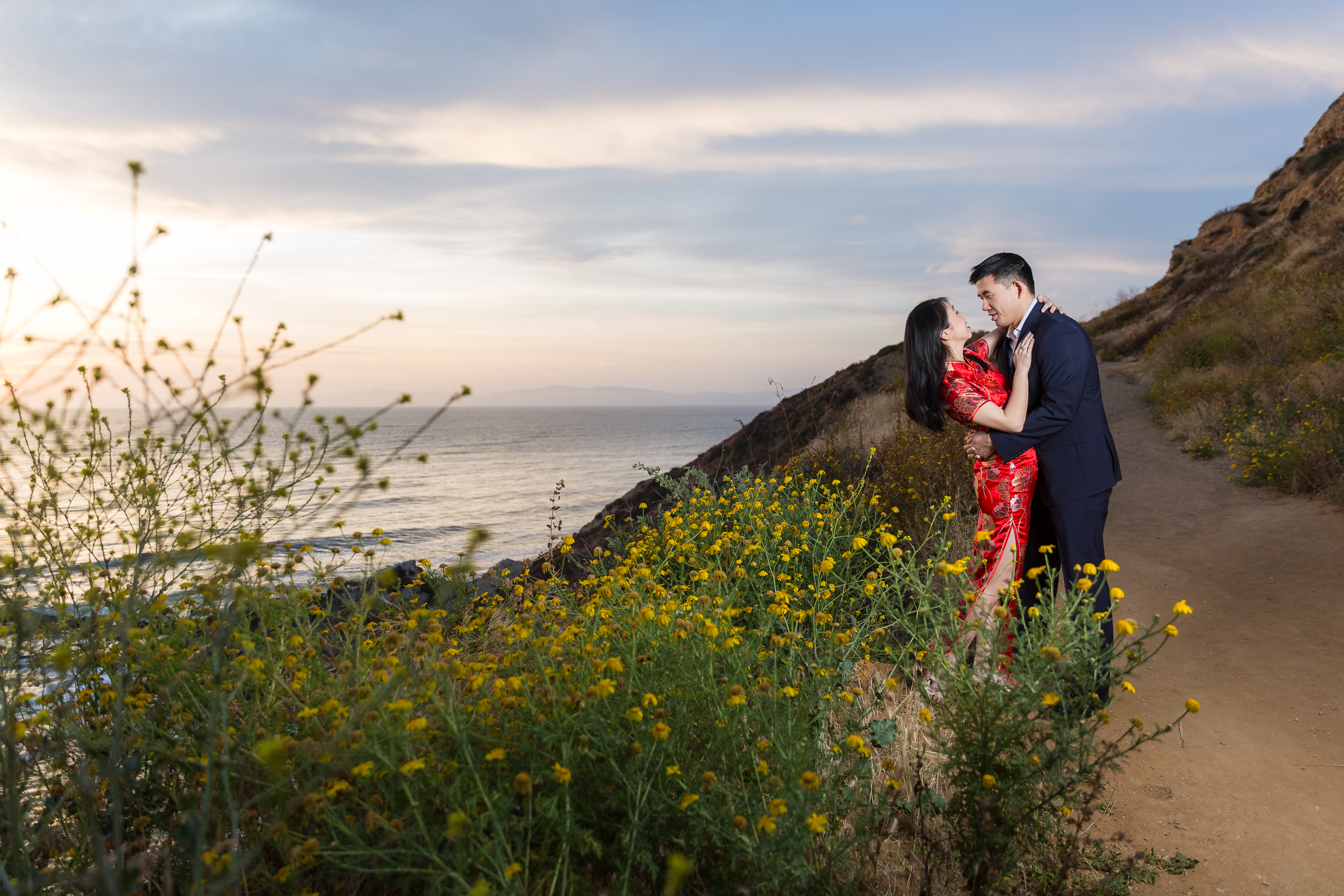 Man dipping woman back behind yellow flowers along cliffside during sunset