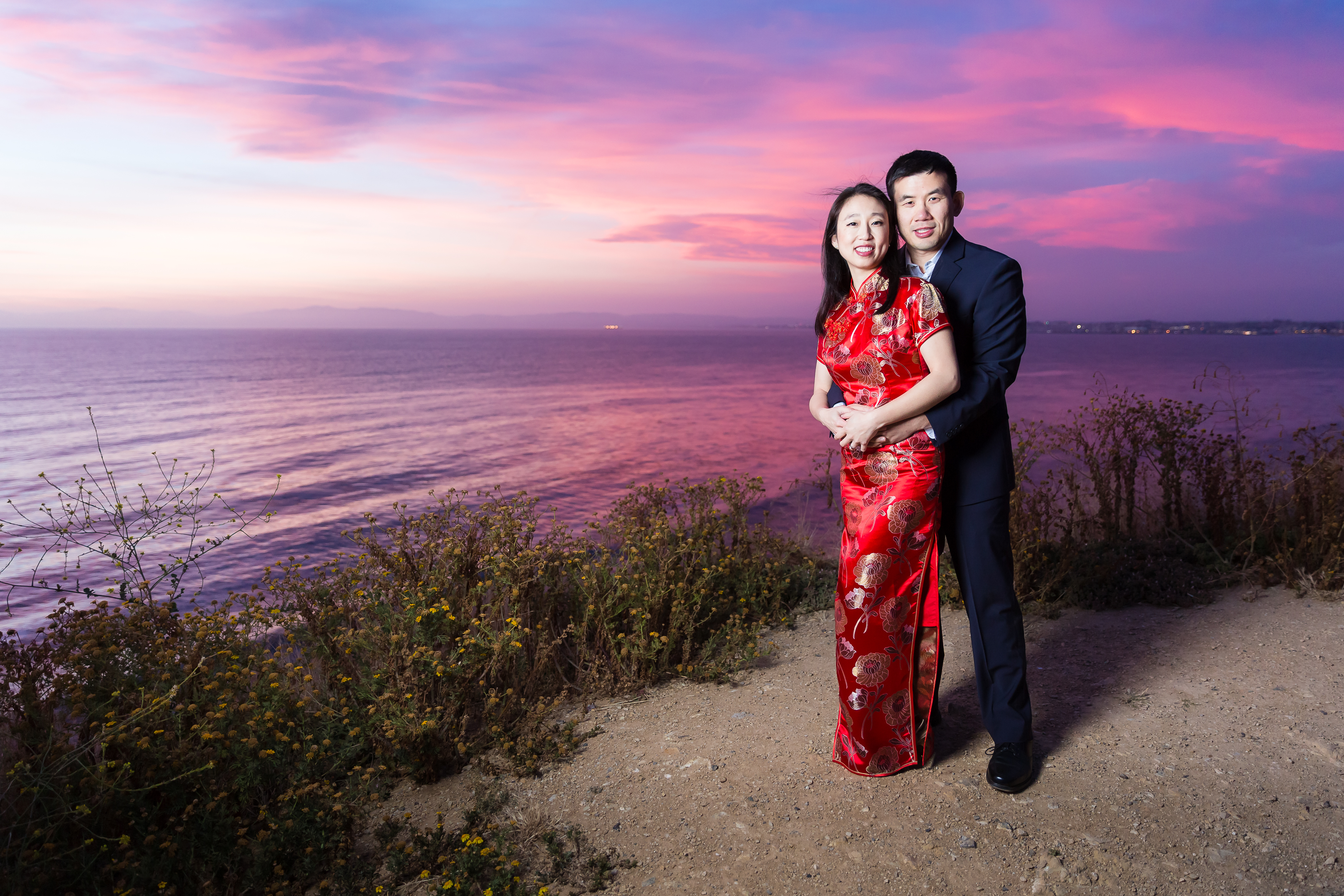 Man hugging woman on cliff edge during pink and purple sunset