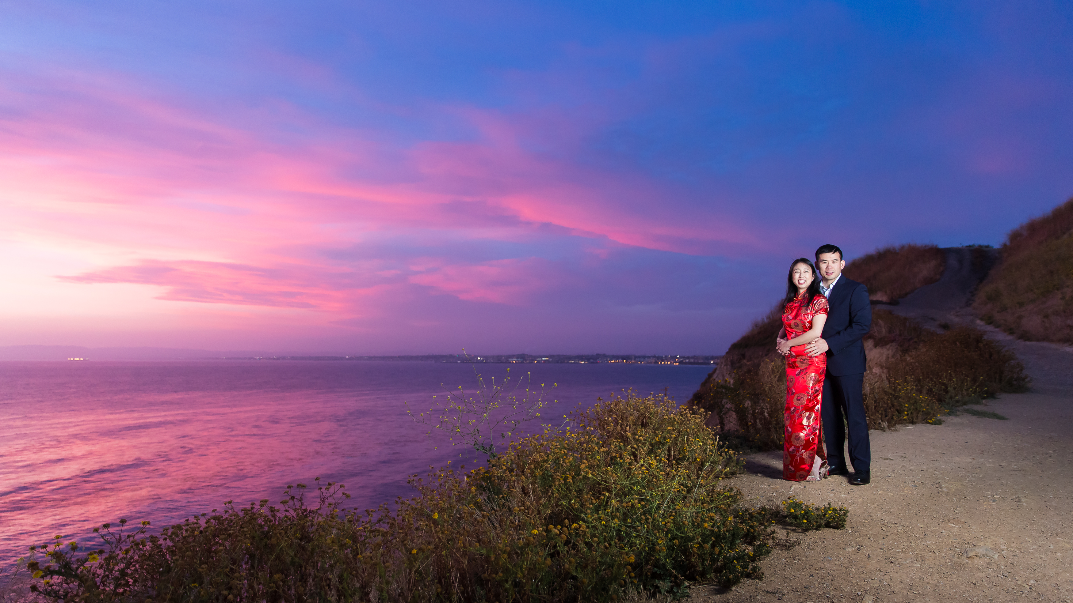 Engaged couple hugging sweetly on cliffside in during epic pink and blue sunset in CA