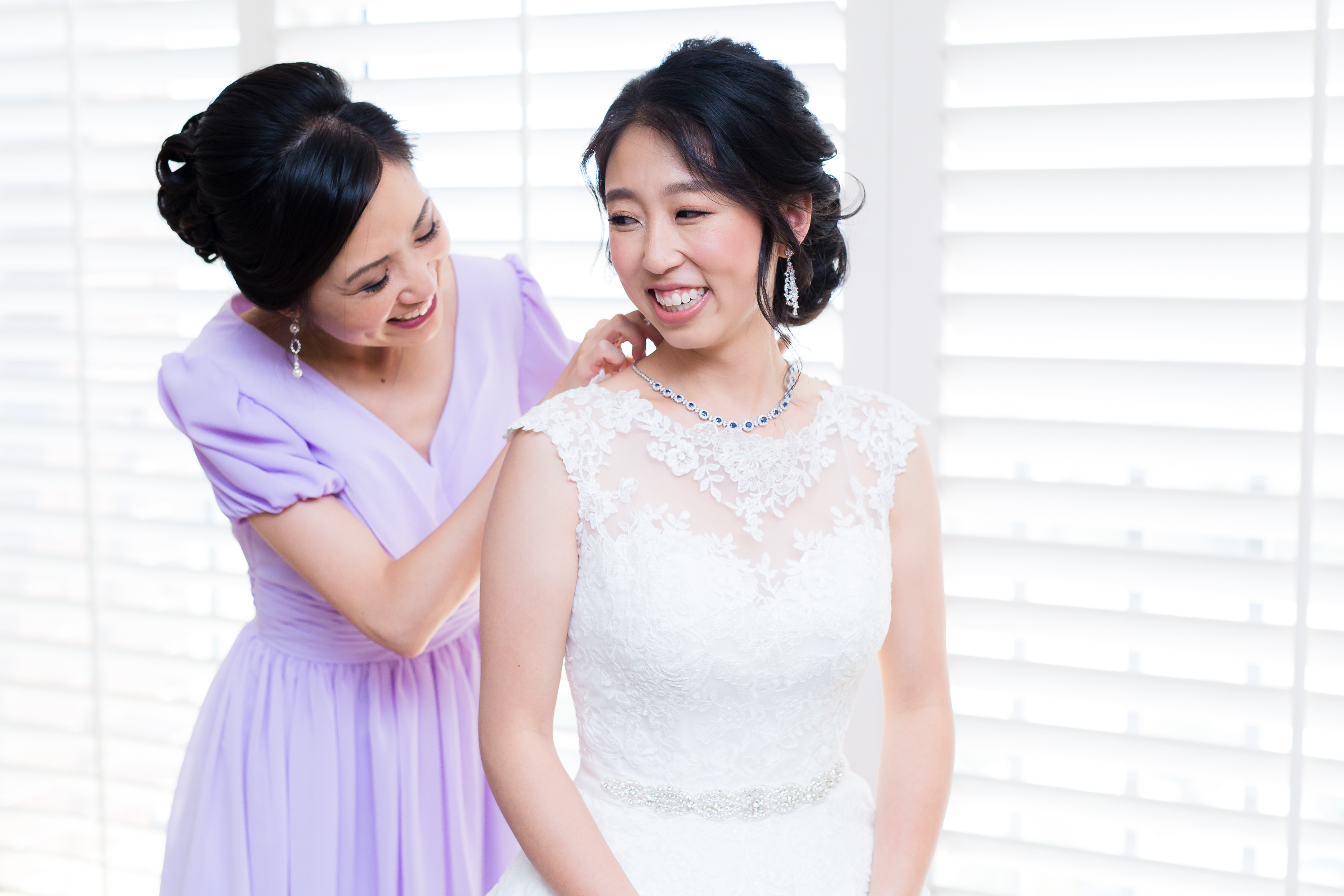 Bridesmaid putting necklace on asian bride next to hotel window