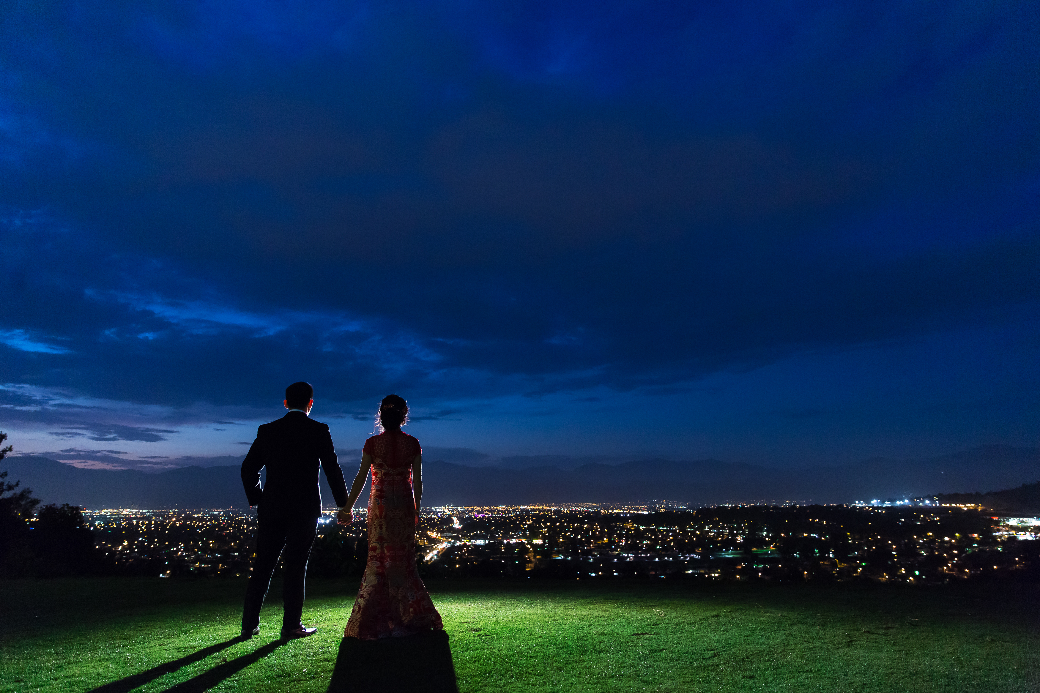 Couple standing on grass hill looking out over city at nighttime