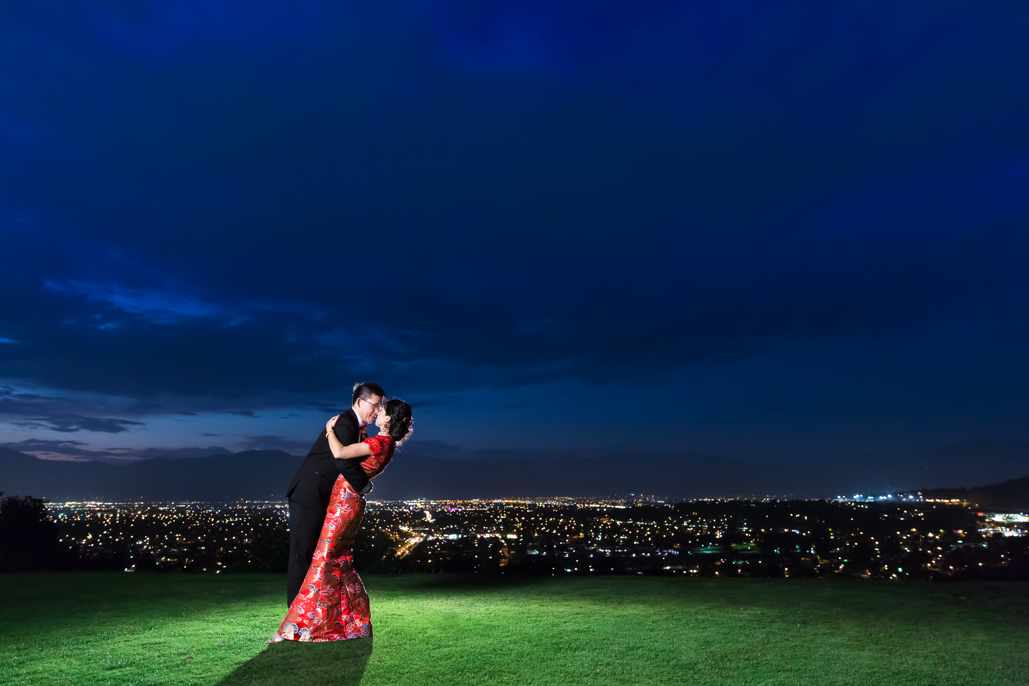 Groom dipping bride for kiss overlooking nighttime cityscape 