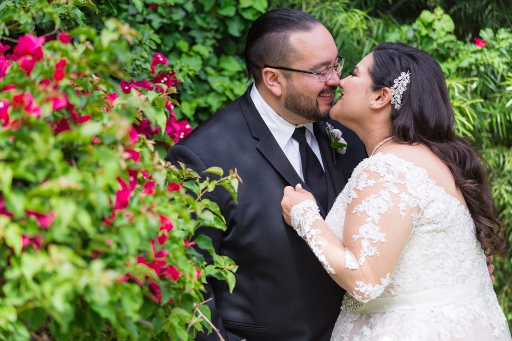Bride and groom smile kissing surrounded by lush greenery and pink flowers