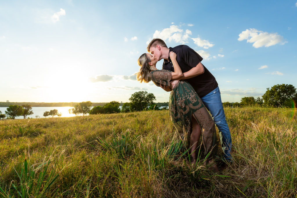 man dipping and kissing woman on grassy field overlooking lake during golden hour engagement session at Dallas Photoshoot Location white rock lake