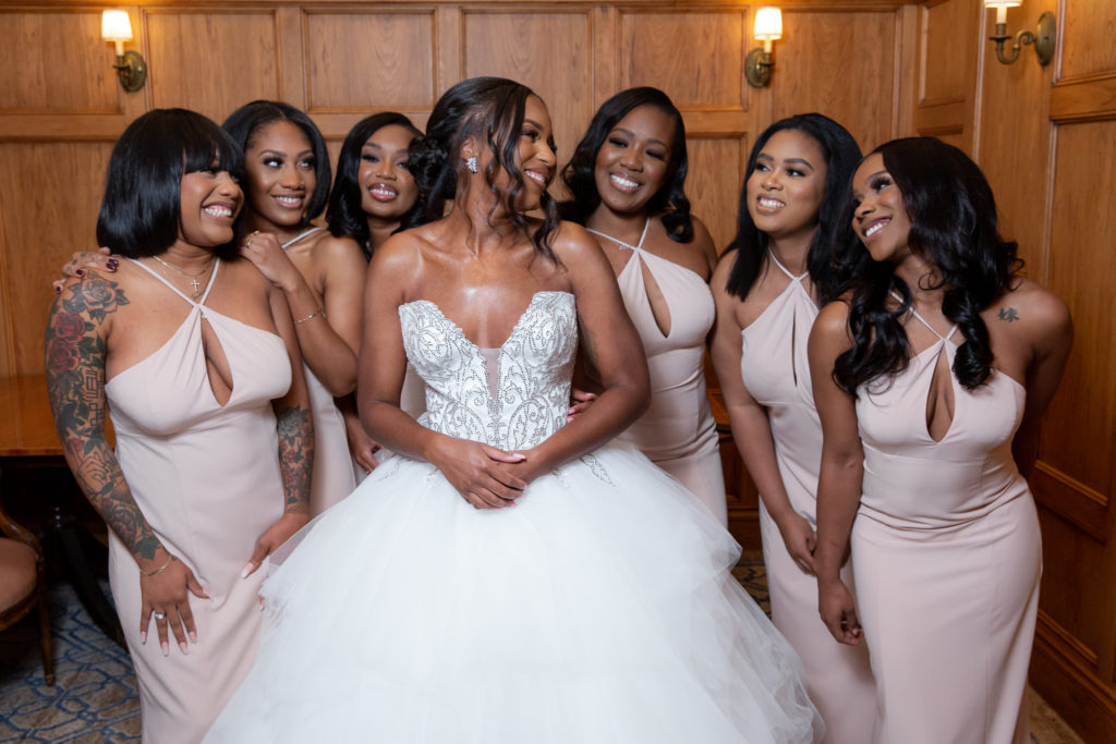 bride surrounded by her bridesmaids who are wearing light pink bridesmaids dresses that are floor length as they smile and laugh together