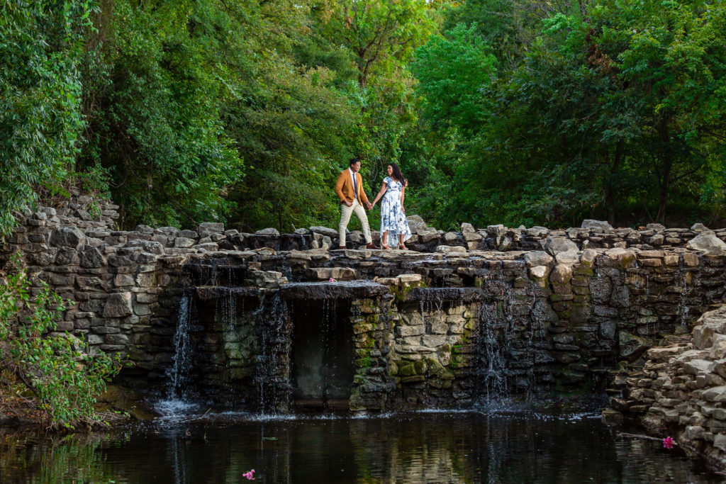 Couple holding hands and walking across waterfall during Prairie Creek Park Engagement Session in Dallas Texas
