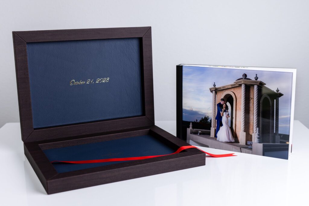 an heirloom wedding album with black spine and an acrylic photo cover of bride and groom hugging on stoney ridge villa wedding venue's rooftop sitting next to it's open luxury dark wood and blue leather box with gold foil and a red ribbon