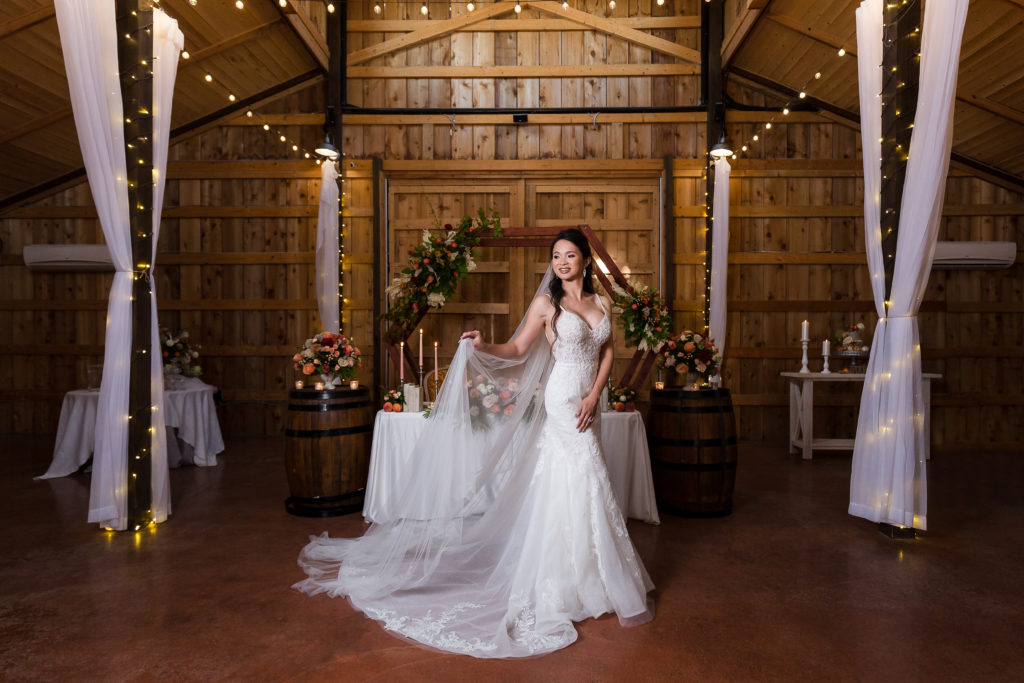 A bride holds her veil in front of her inside barn decorated in twinkle lights for a wedding reception at the big white barn in Decatur TX