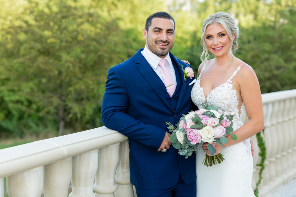 Outdoor wedding photo with bride and groom standing together on an outdoor patio with the groom in a dark blue suit leaning against a marble railing and his bride leaning into him while holding a pink floral bouquet at Montclair wedding venue in Colleyville