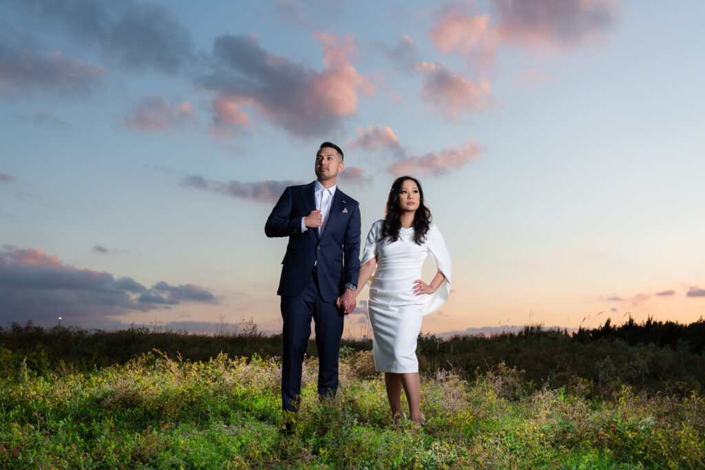 engagement photographer dallas captures man and woman holding hands in a field with a sunset behind them as they are dressed in formal outfits with man in a blue suit and woman in a white cocktail dress