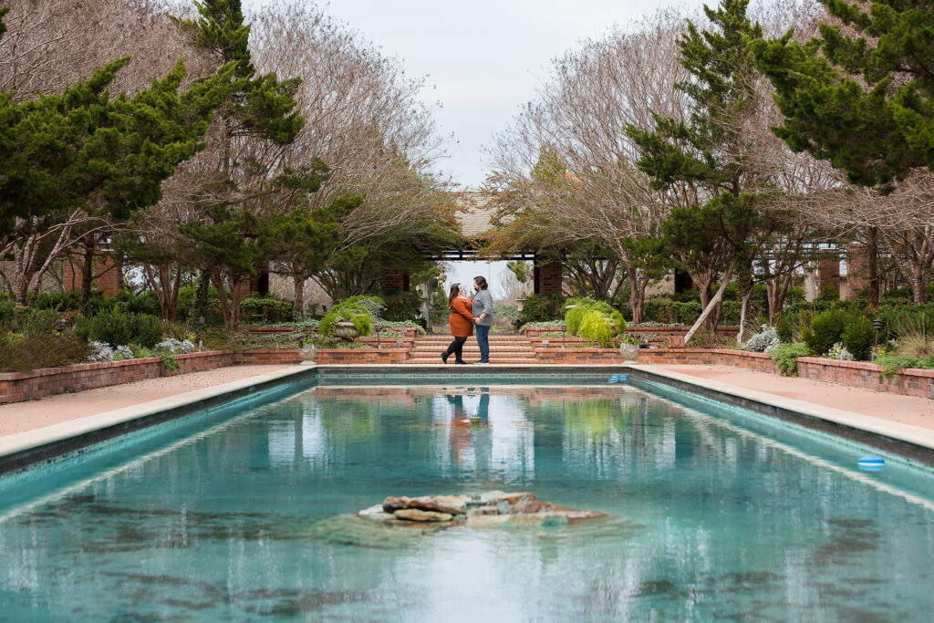 dallas wedding photographer captures dfw engagement pictures with man and woman holding hands at the end of a reflective pool in Clark Gardens in weatherford
