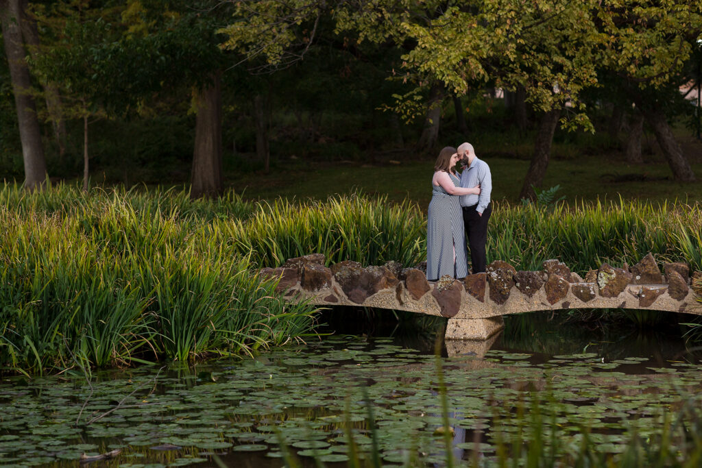 Dallas wedding photographer captures engagement pictures at Texas Women's University with woman leaning onto a man as they stand together on a bridge over a lake filled with Lilly pads for engagement session
