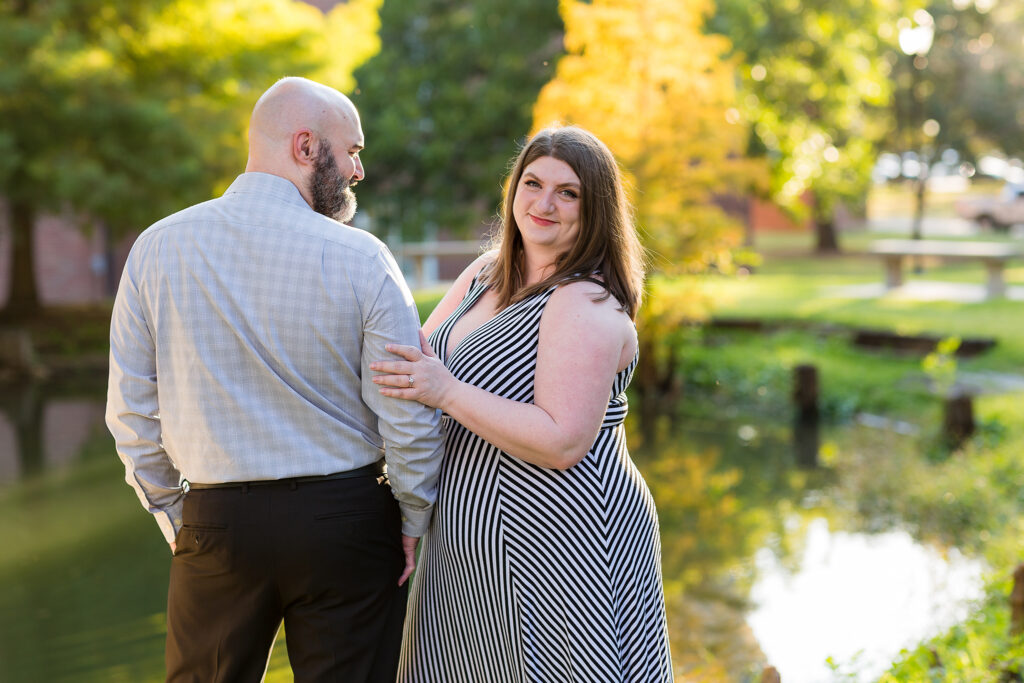 Man facing lake while woman snuggling into his arm smiling at the camera with a lake and trees behind them for engagement photos on TWU campus