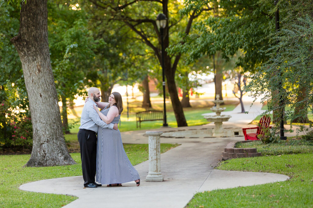 Texas Women's University engagement pictures with man and woman in a courtyard together as the woman holds onto the mans shoulder and they smile to one another with a path lined by trees is behind them captured by Dallas wedding photographer