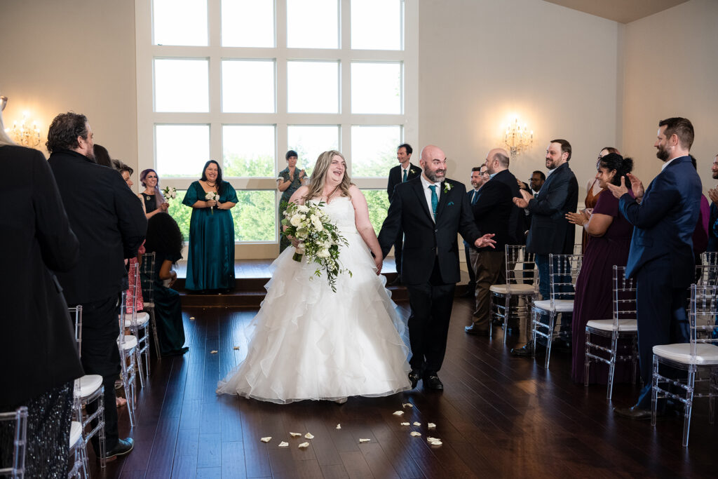 Bride and groom walk back down the aisle smiling and waving at friends and family at milestone denton wedding venue