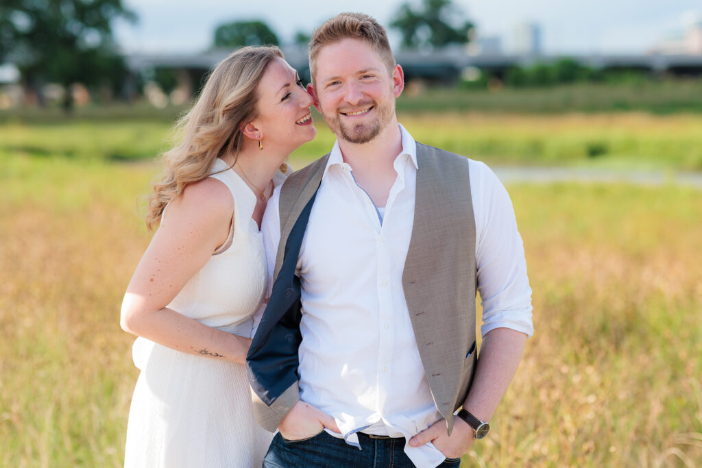 Engaged woman whispers into fiancé's ear during engagement session at Trinity Overlook Park in Dallas TX by Stefani Ciotti Photography
