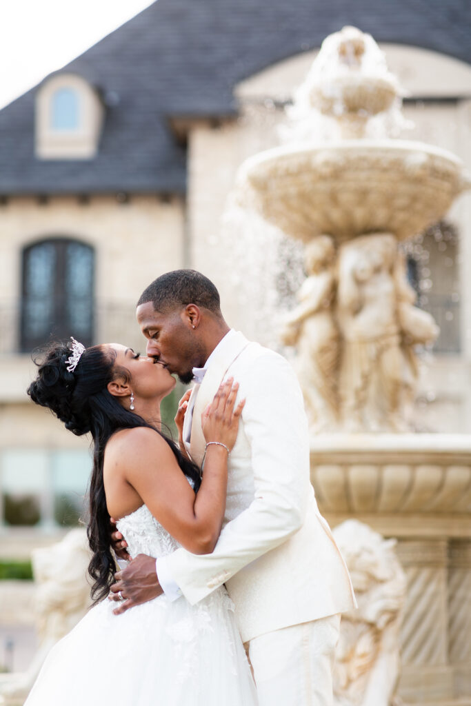 Dallas wedding photographer captures couple kissing in front of fountain on knotting hill place wedding venue front lawn in little elm texas