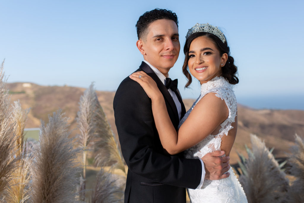 dallas wedding photographer captures bride and groom overlooking mountains and ocean with black tux and lace white wedding dress while wearing a bridal crown 