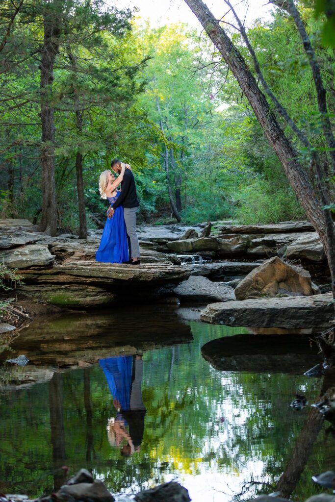 woman in blue dress hugging fiancé on sheet rock above still river surrounded by trees at stone creek park in flower mound texas during engagement session