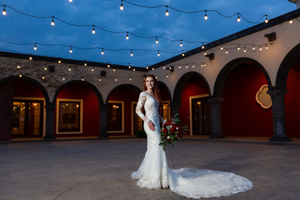outdoor bridal pictures at night with bride in a lace wedding dress with a large train standing on an outdoor patio with string lights over head photographed by Dallas wedding photographers