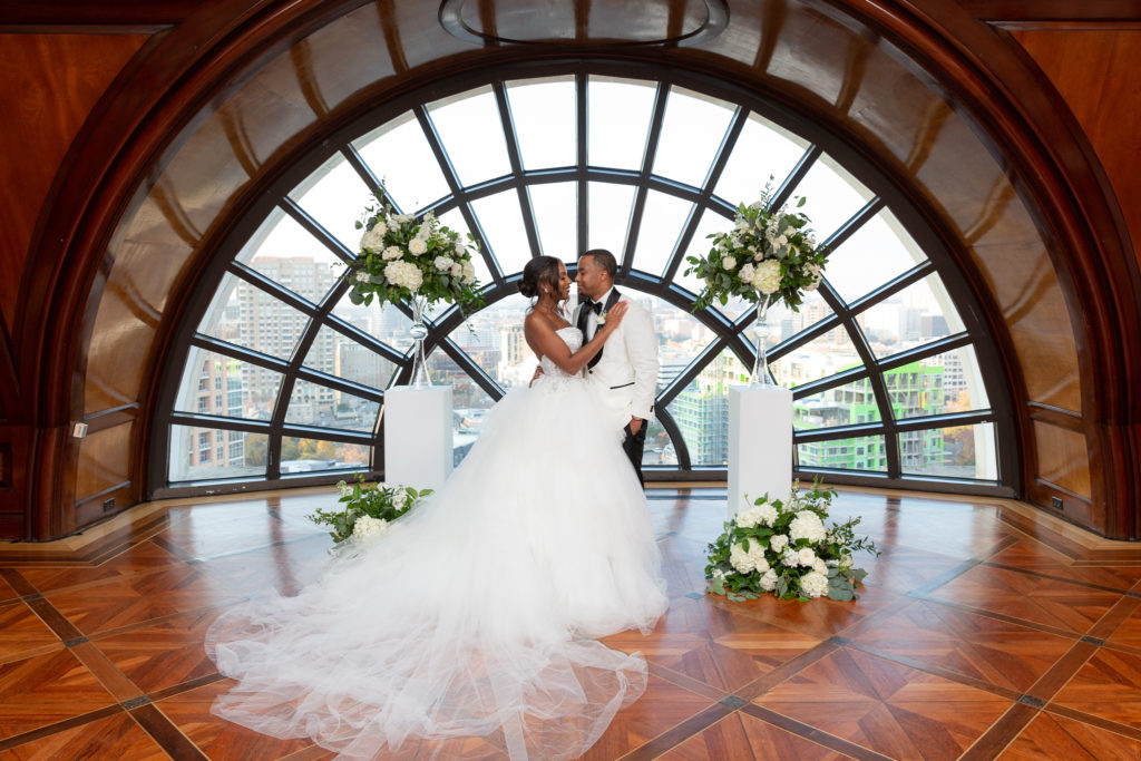 bride and groom in a luxurious ballroom at Hotel Crescent Court wedding venue standing in front of a hald moon window with white florals surrounding them as they embrace captured by dallas wedding photographers