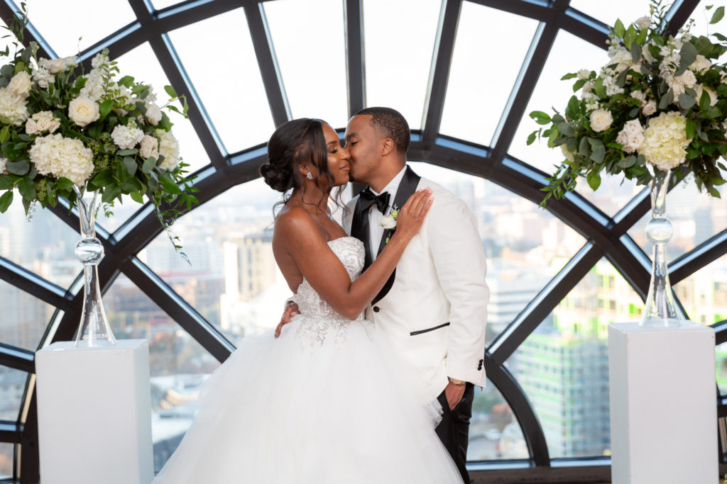 bridal portraits with bride and groom kissing in front of a large window at the top of a Dallas hotel photographed by Dallas wedding photographers