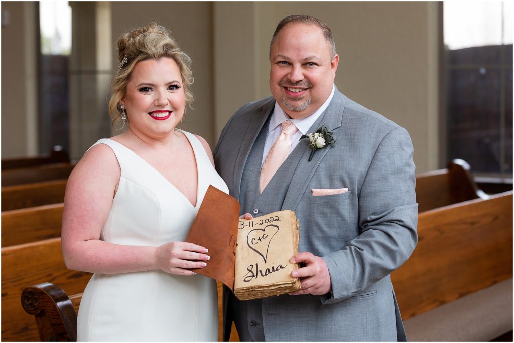 Bride and groom stand side by side displaying a leather and rough paper guestbook with a heart on the front page captured by Dallas wedding photographers