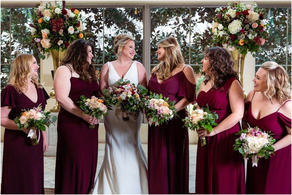 Bride laughs with five bridesmaids in front of floral arrangements at Ana Villa TX chapel by Dallas wedding photographers