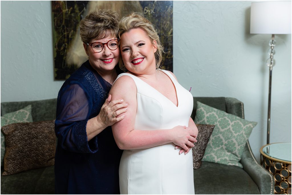 Dallas wedding photographers capture the bride's mother and the bride posing and smiling together in front of a green velvet sofa at The Colony TX