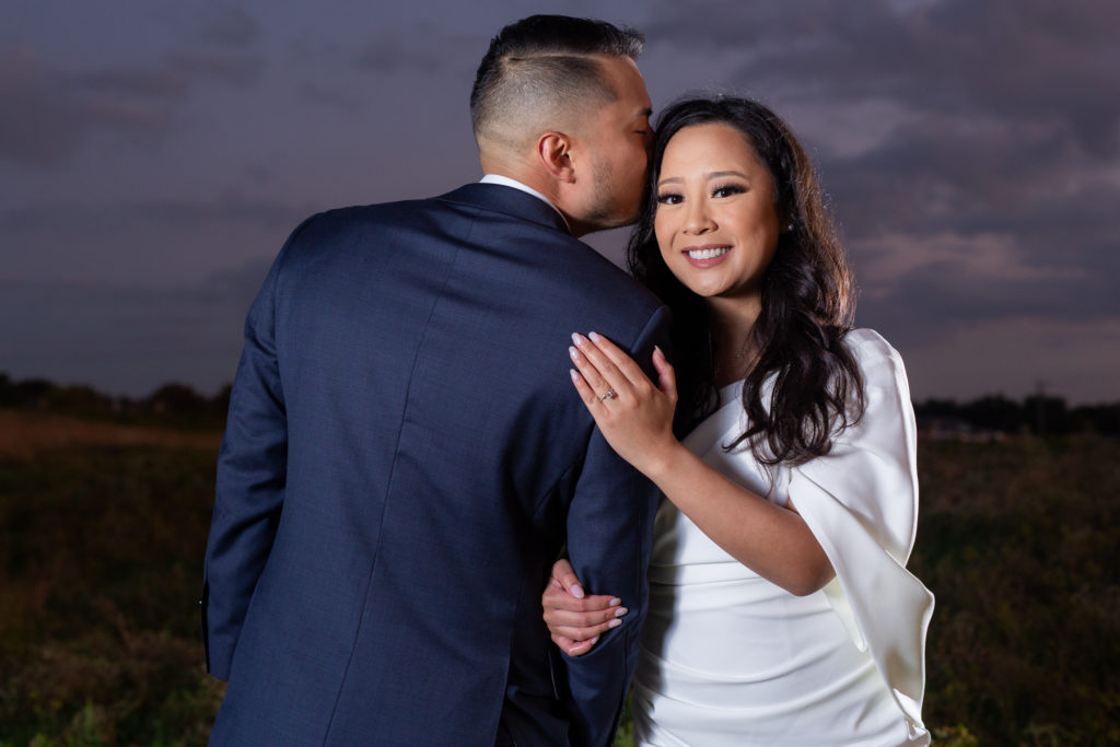 Groom kissing bride on temple while bride shows off engagement ring