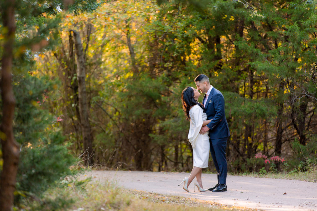 Plano park engagement session with woman leaning into the man and kissing him as her foot pops up and man leans into the woman as they stand on a path together