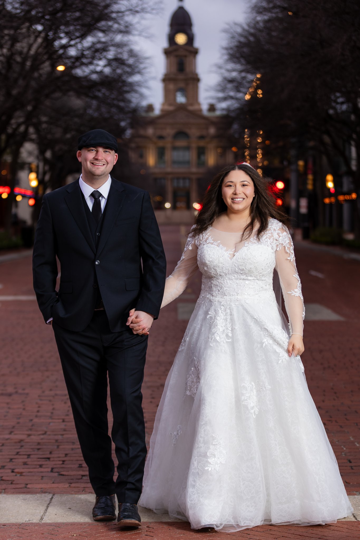 Wedding couple smiling, holding hands and walking down Sundance Square in downtown Fort Worth with Tarrant County Courthouse in background