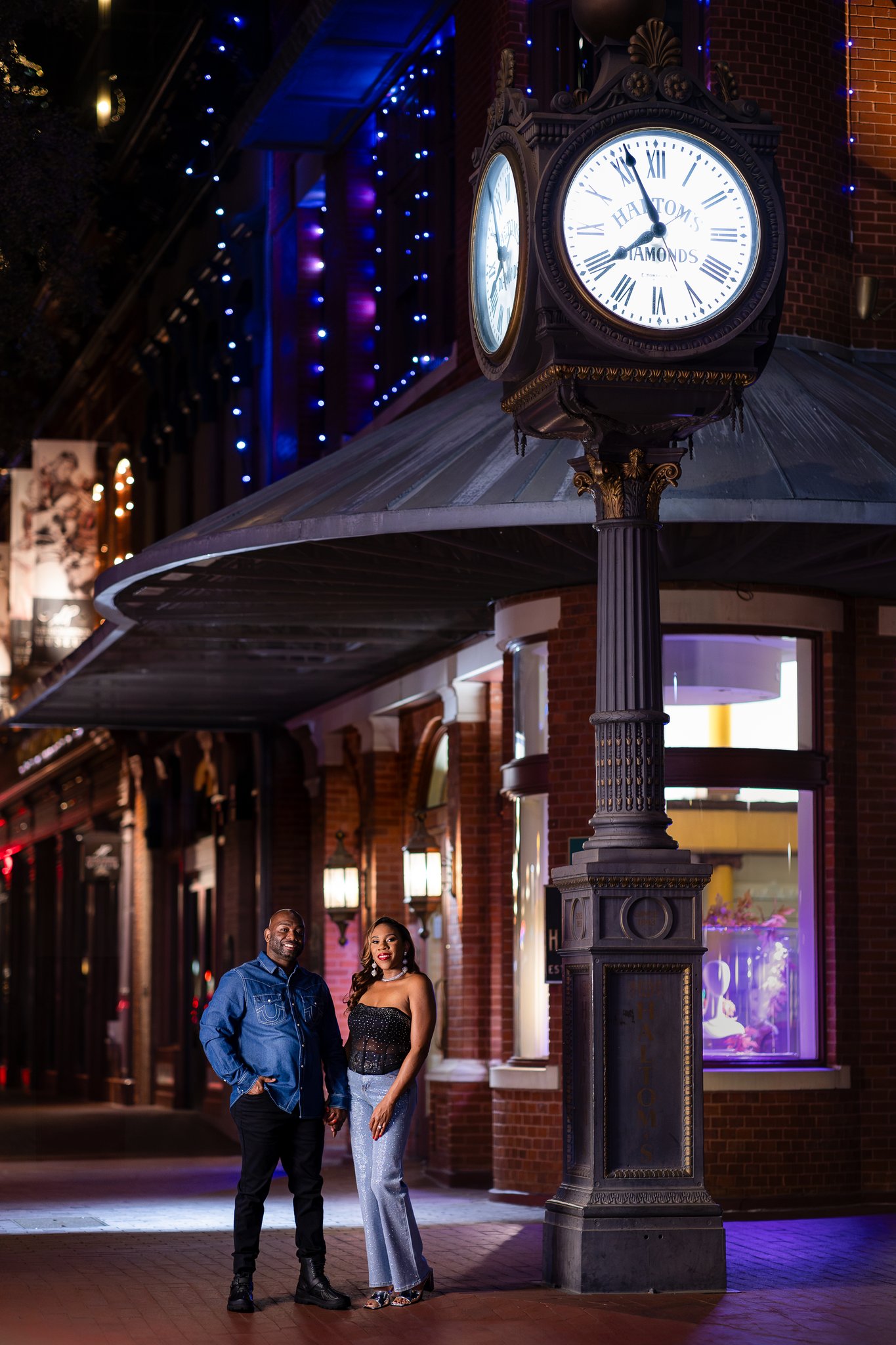 Engaged couple standing together at night by big clock near Sundance Square in downtown Fort Worth during engagement session