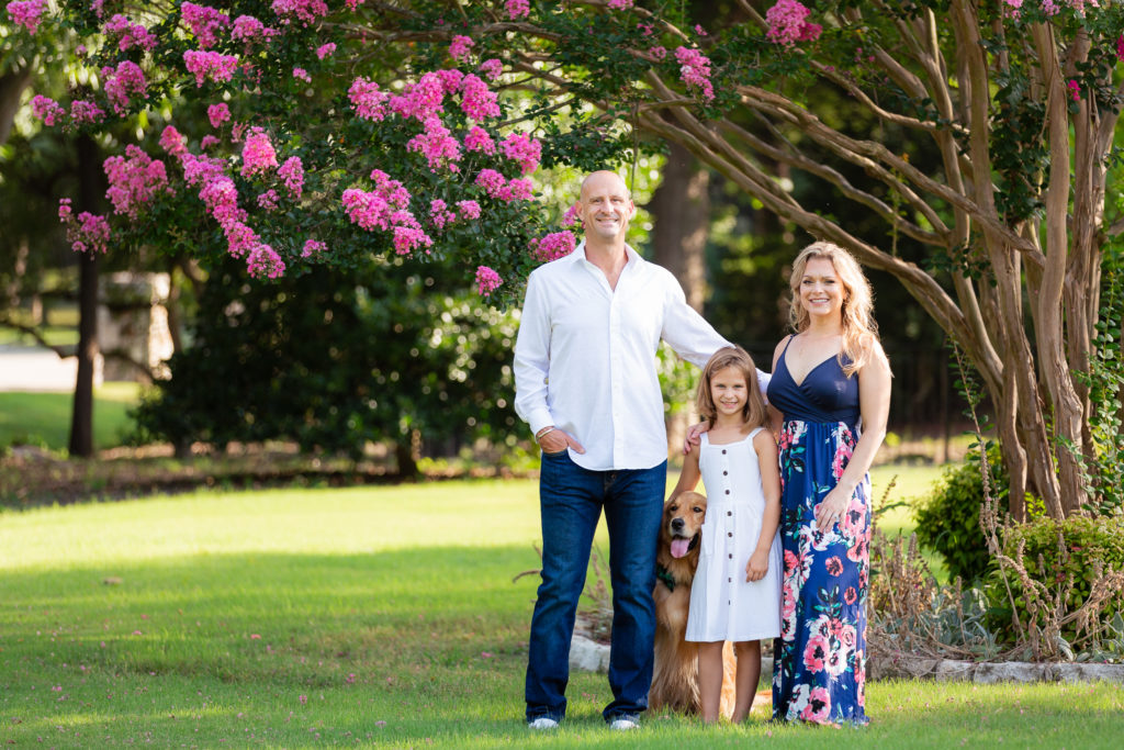 dallas wedding photographers captures Adriatica Village Photos For Families with mother and father standing with their young daughter and dog under a pink floral tree for summer family photos