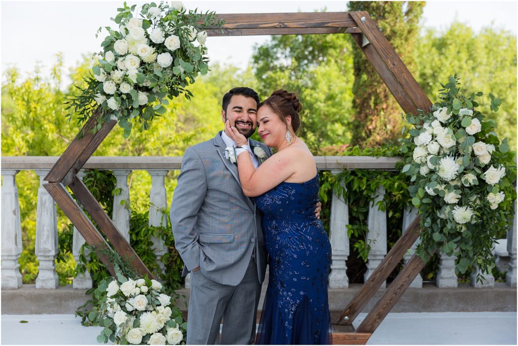 groom posing with his mother as she holds his face and leans into his while they stand in front of a wedding arch decorated with white florals and greenery at a Dallas wedding venue captured by Dallas wedding photographers