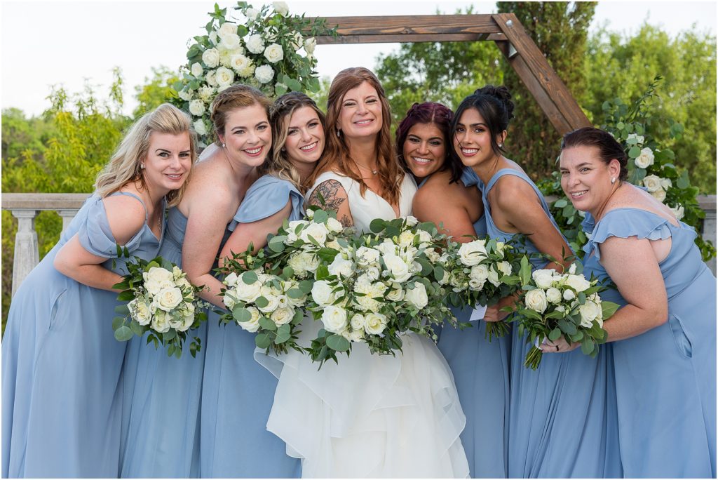 bride standing with her bridesmaids as they all hold their white floral bouquets together and smile while leaning into each other captured by Dallas wedding photographers