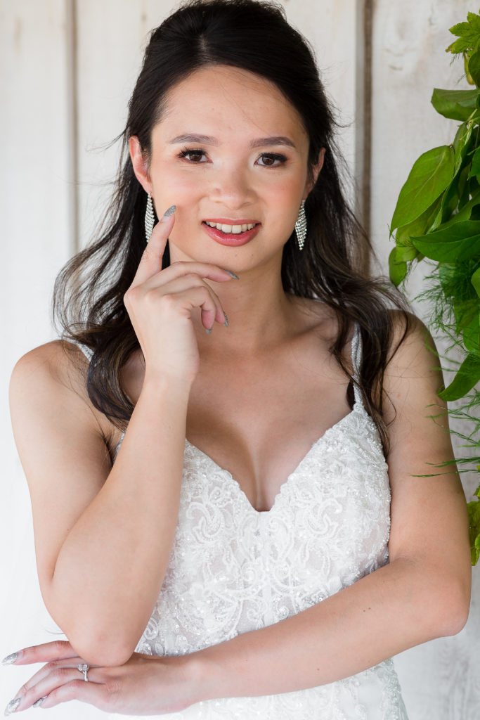 A bride in a lace sweetheart neckline dress poses and smiles in front of a white barn door for Dallas weddings photographers at a venue in Dallas TX