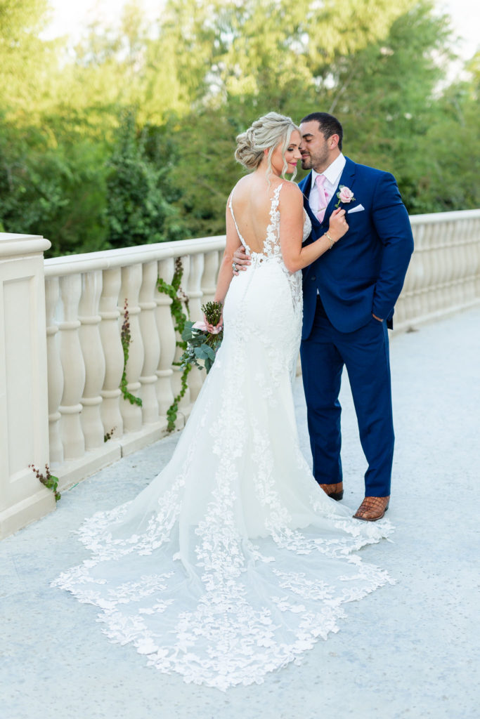Outdoor wedding photos with bride and groom on a large deck with marble floors with bride standing in front of her groom and holding his tux while her long lace wedding dress trails behind her and her gremlins in to kiss her captured by Dallas wedding photographer