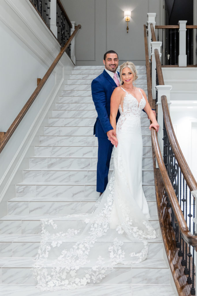 Dallas wedding photographer photographs bride and groom standing on a marble staircase inside of their Dallas wedding venue with groom standing behind his bride and holding her hand as her train falls down the stairs