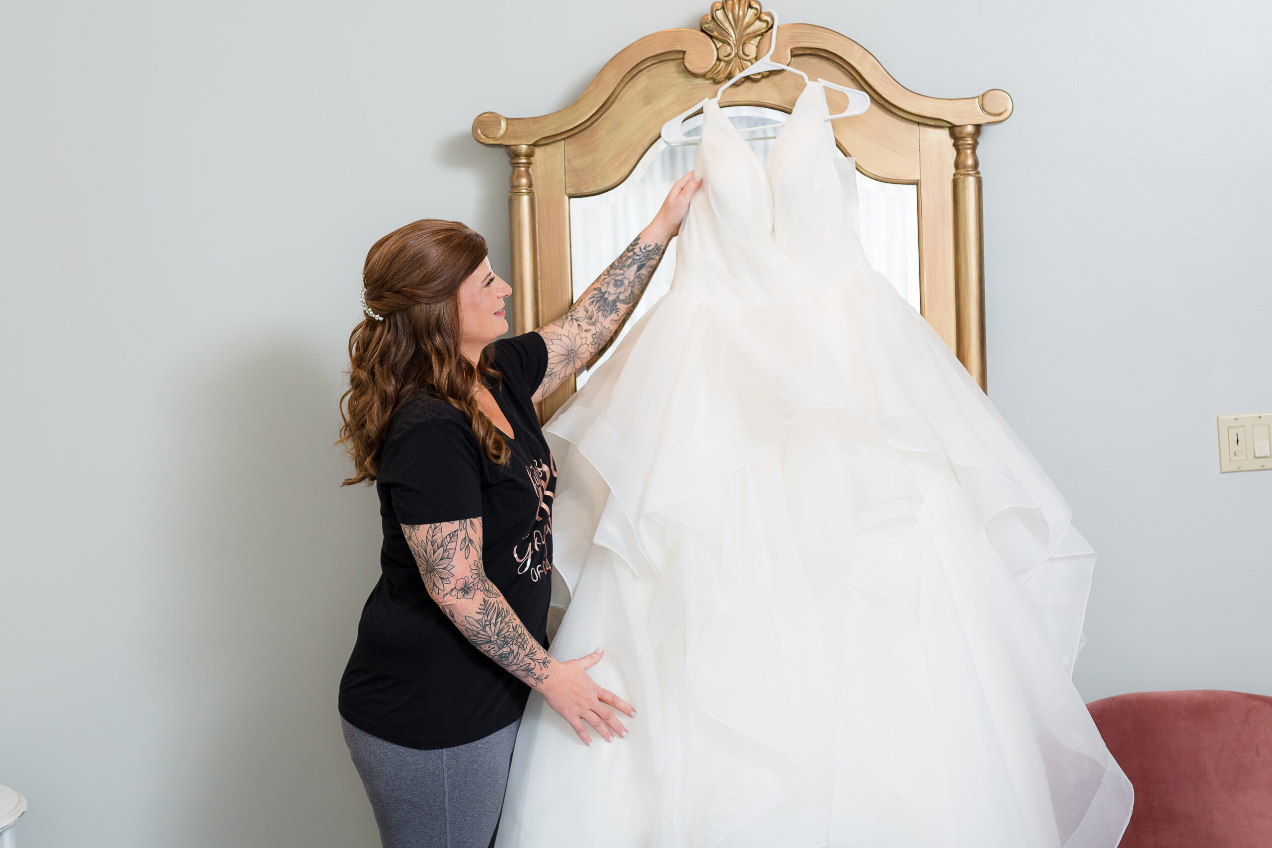Bride picking up her satin wedding dress that is hanging on a white hanger on a gold mirror the morning of her wedding captured by dallas wedding photographers