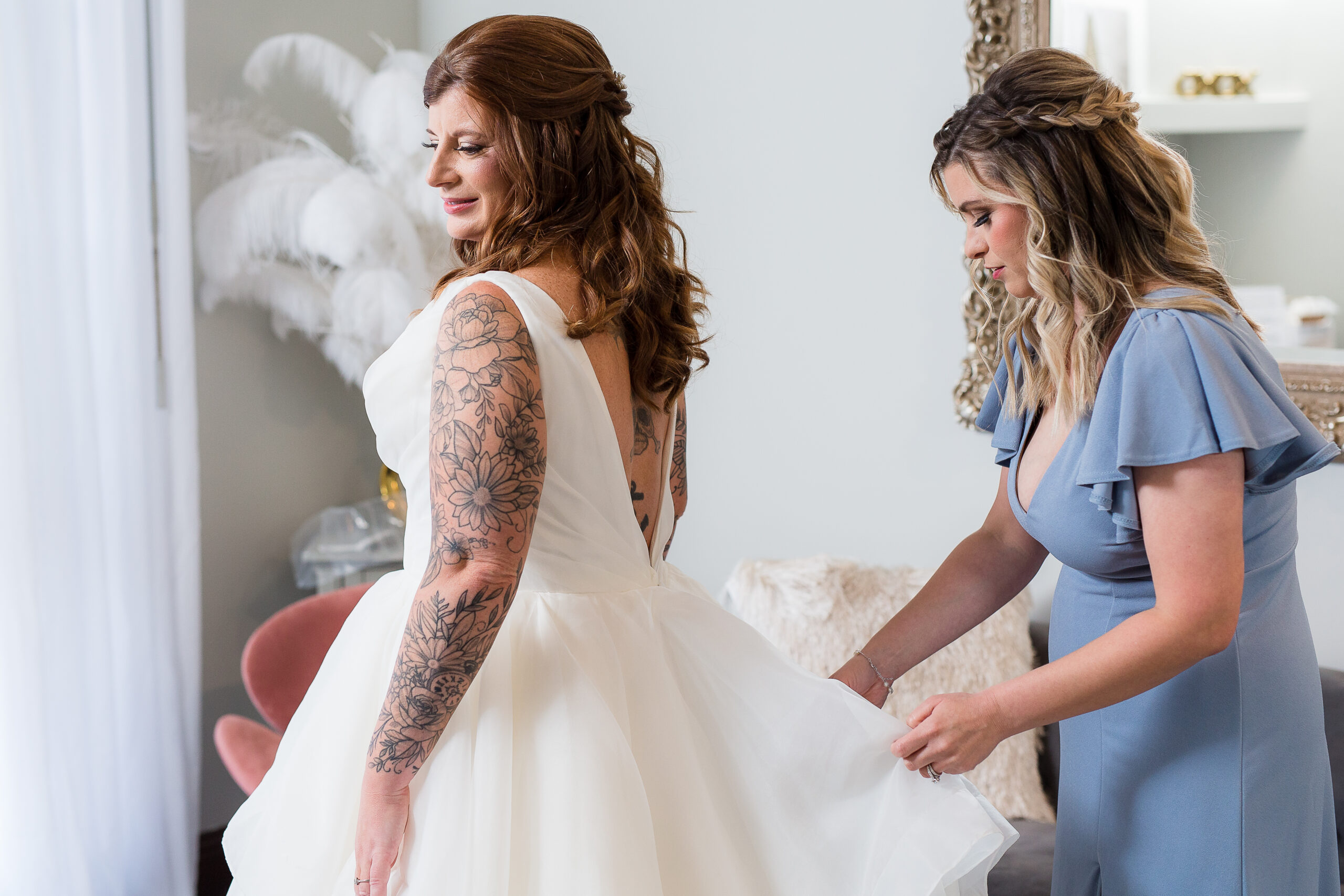 The morning of the wedding bride gets into her wedding dress as her maid of honor who is in a dusty blue bridesmaid stress helps her get ready