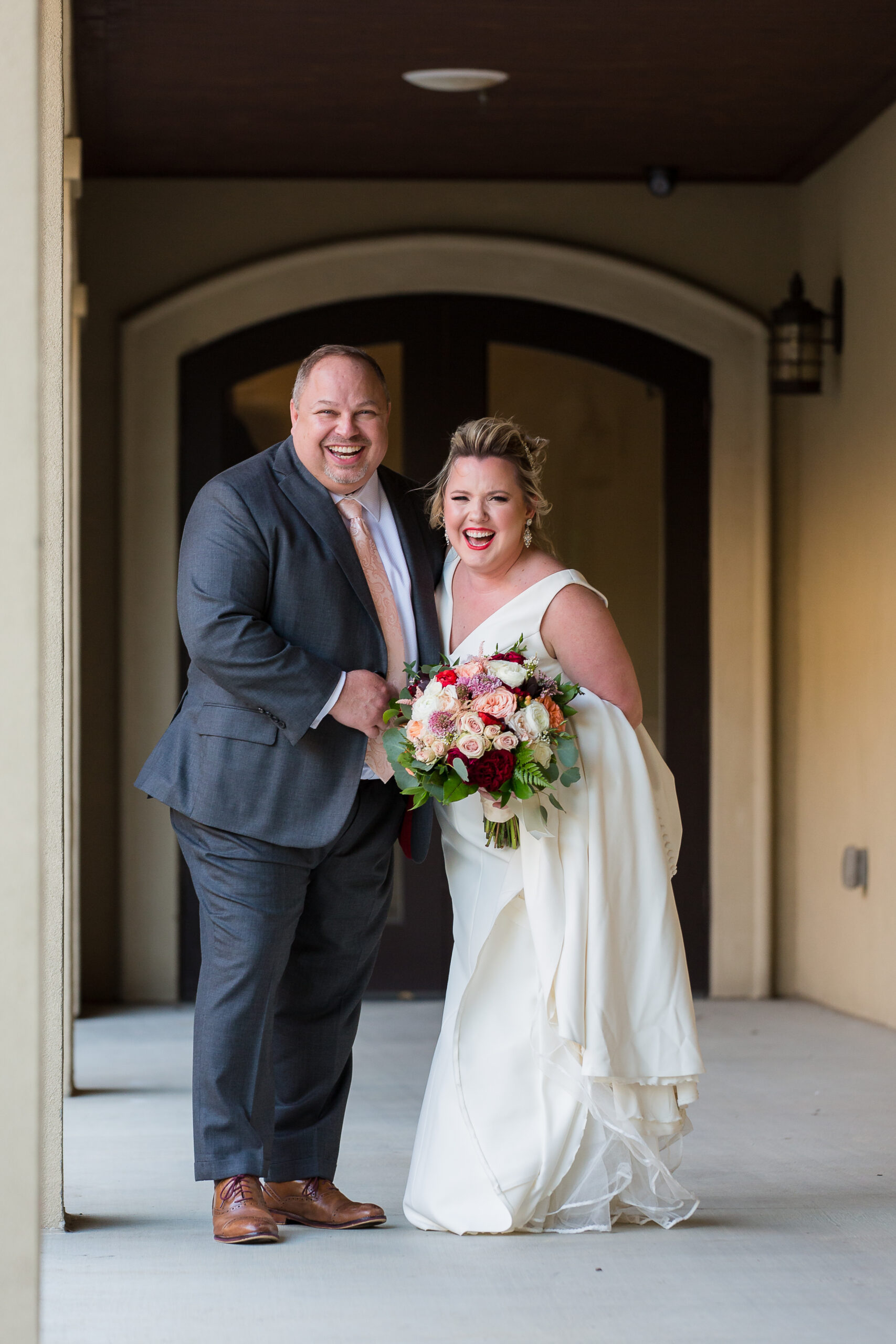 Dallas wedding photographer captures bride and groom walking down a corridor together together with the bride holding her train and a bouquet as they look over their shoulders laughing for their Dallas wedding day