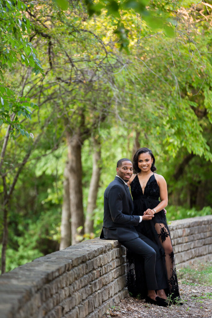 Dallas wedding photographers captures outdoor session with man sitting on a stone half wall with his fiance standing next to him and smiling as they hold hands for engagement photos dallas