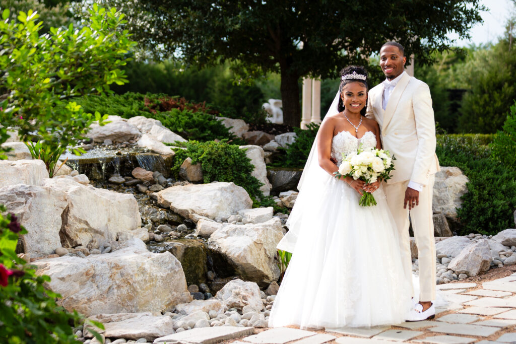 Bride and groom in white smiling at camera in front of lush rock waterfall at Knotting Hill Place wedding venue in Little Elm Texas