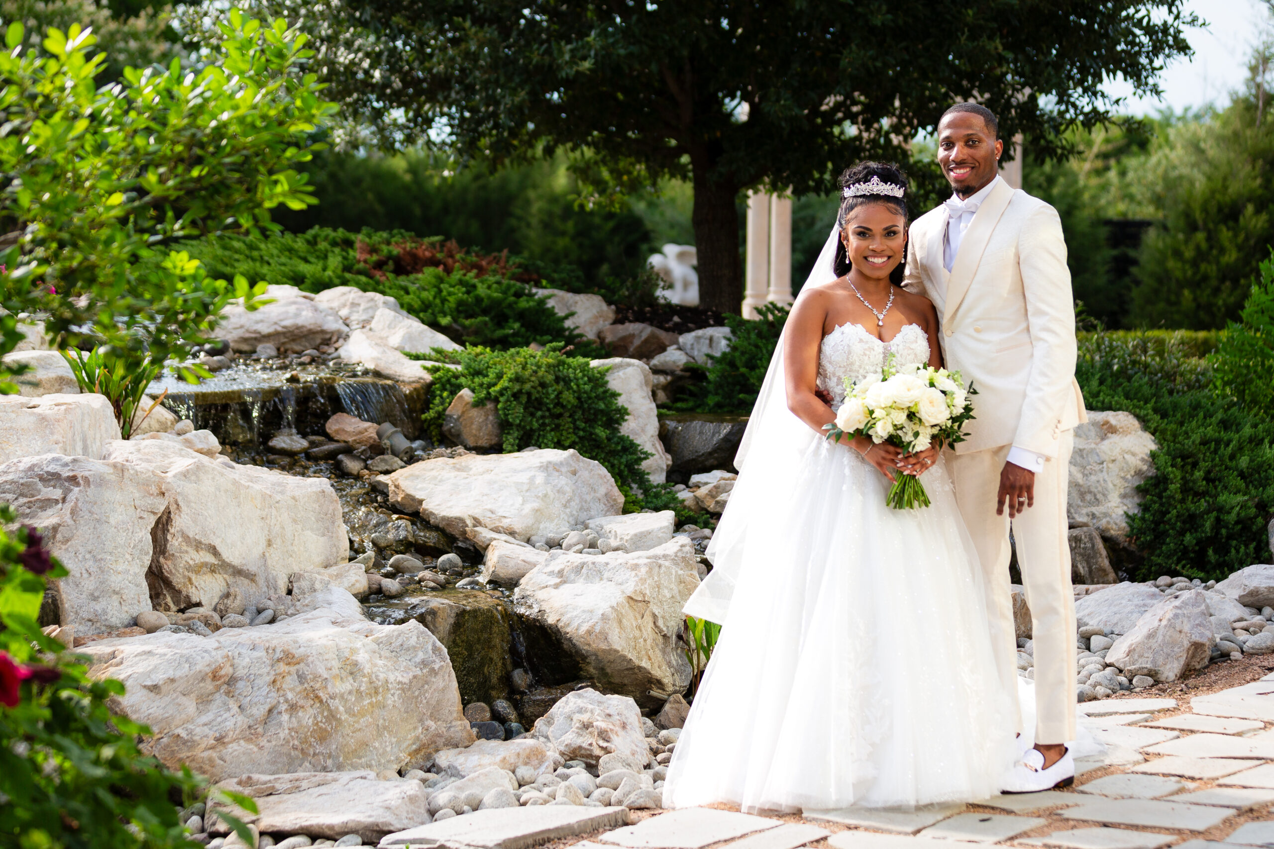 Bride and groom dressed in all white standing together in front of lush waterfall feature at Knotting Hill Place wedding venue in Little Elm Texas