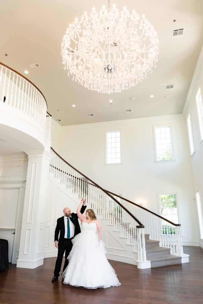 Bride and groom dance by a grand staircase in the beautiful entry way of the Milestone Denton wedding venue under an impressive chandelier.