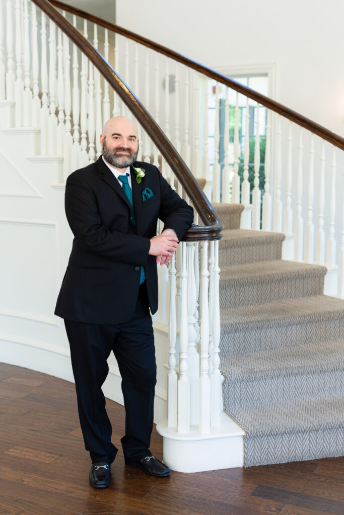 Groom standing against staircase in black suit and teal tie before wedding ceremony